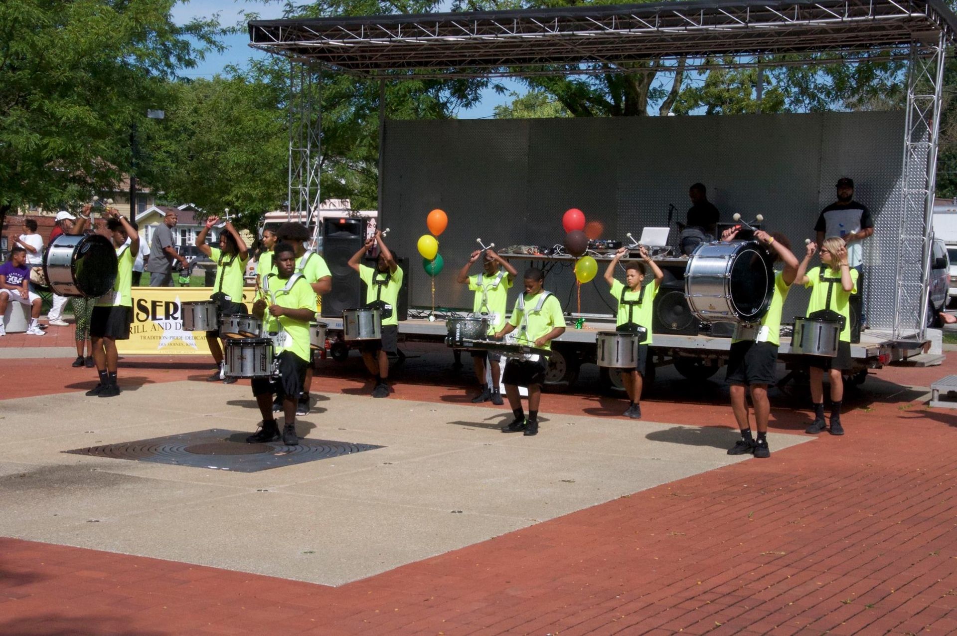 Drumline performing on an outdoor stage with colorful balloons and people watching in the background.