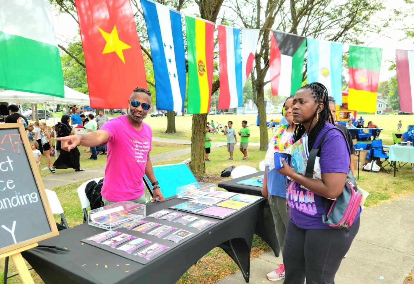 Outdoor multicultural event with flags displayed, people at a booth discussing and sharing materials.